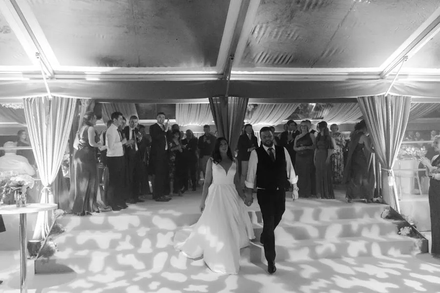 Bride and groom walking hand in hand into the marquee as guests applaud, in black and white