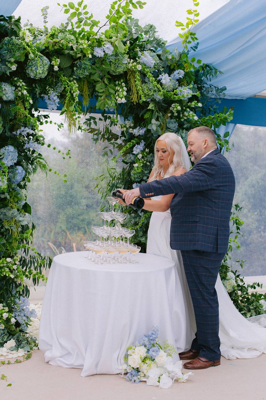 Couple pouring champagne over a glass tower beside a flower arch