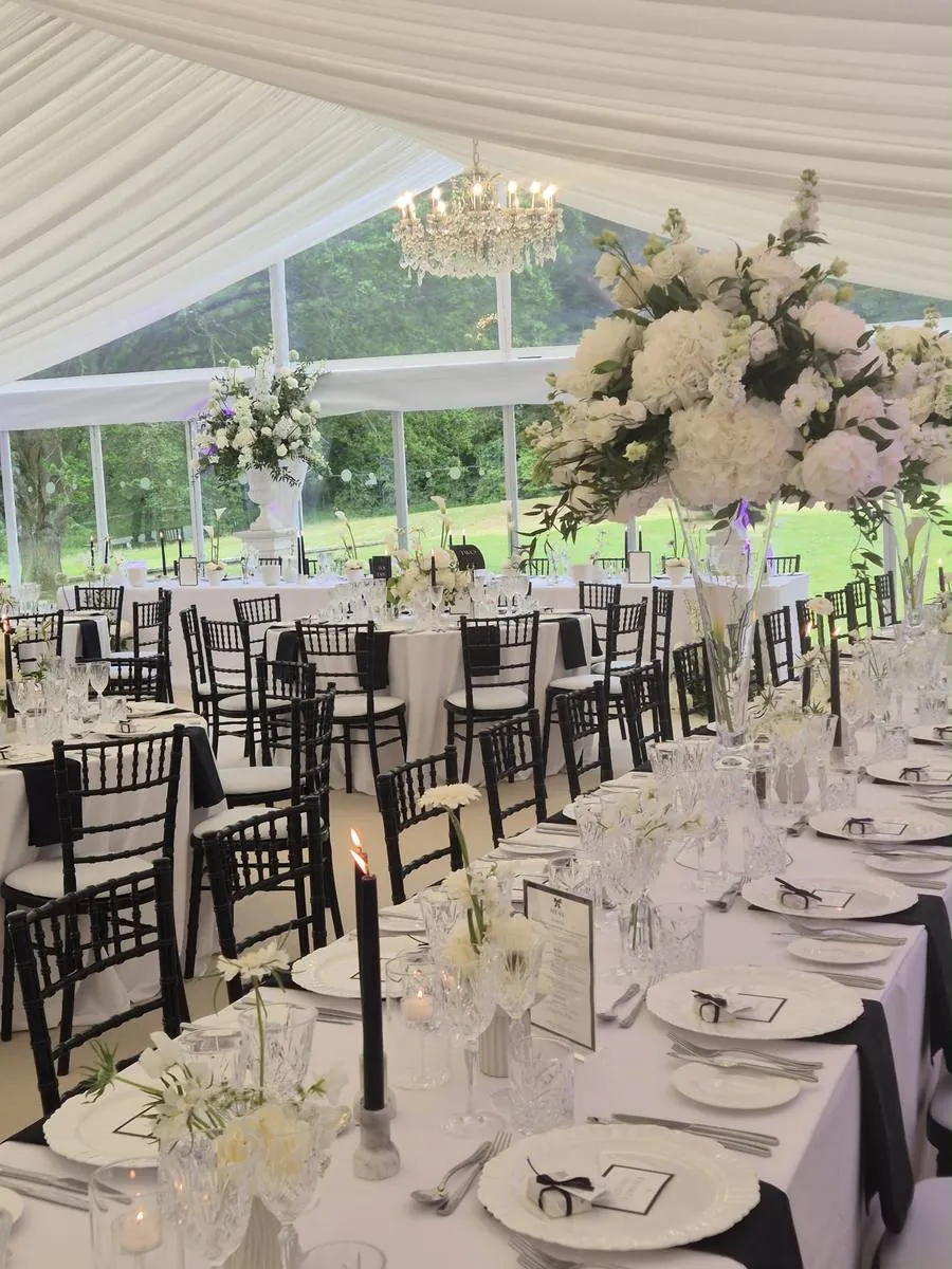 Marquee dining with black chairs, white linen and tall white floral centrepieces